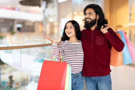Foto de Emocionada joven pareja hindú de pie en el centro comercial, abrazando y sonriendo, apuntando al espacio de copia en blanco, sosteniendo coloridas compras de bolsas de papel, disfrutando de compras de venta de temporada juntos - Imagen libre de derechos
