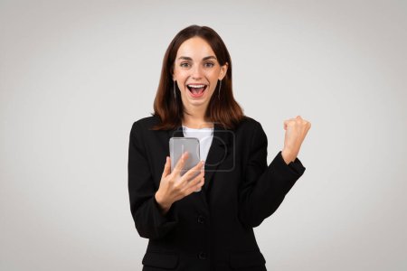 Foto de Emocionada empresaria europea milenaria con una expresión alegre, mirando su teléfono inteligente y celebrando el éxito con una bomba de puño, vestida con un traje negro profesional - Imagen libre de derechos