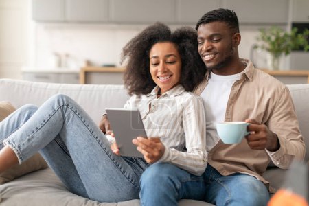 Téléchargez les photos : Sourire jeune couple noir relaxant avec tablette sur canapé intérieur, profiter week-end numérique ensemble tout en regardant un film en ligne, la navigation et le partage de moments en ligne - en image libre de droit