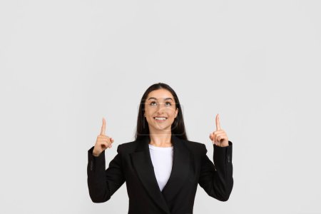 Téléchargez les photos : Femme professionnelle souriante en costume noir pointant vers le haut avec les deux doigts à l'espace libre, exprimant la positivité et la direction vers quelque chose au-dessus - en image libre de droit