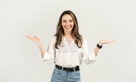 Foto de Una joven europea animada con el pelo moreno, vistiendo una blusa blanca y jeans, sonriendo y sosteniendo sus manos en un gesto de equilibrio, sobre un fondo claro, estudio - Imagen libre de derechos