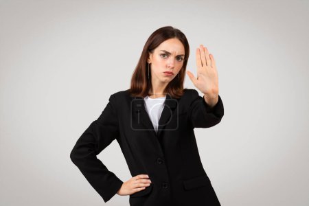 Photo for Stern millennial european businesswoman showing a stop hand sign with a serious expression, emphasizing boundaries or denial, in a formal black suit on a neutral background - Royalty Free Image