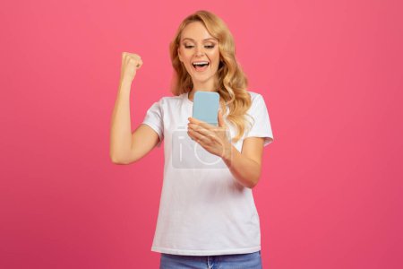 Joyful Blonde Young Woman Holding Smartphone Shaking Fist, Making Yes Gesture In Excitement While Reading Positive Message, Standing Over Pink Studio Background, Celebrating Great News, Success