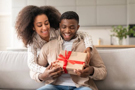 Photo for Young joyful black couple celebrating holidays and special occasions, woman presenting wrapped gift box to husband, sharing joy sitting on couch at home interior. Valentines day present - Royalty Free Image