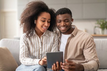 Téléchargez les photos : Couple afro-américain souriant avec écouteurs sans fil utilisant une tablette numérique pour des activités en ligne et du plaisir, immergé dans la technologie gadget, profiter d'un week-end confortable ensemble sur le canapé dans le salon - en image libre de droit