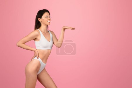 Photo for Vivacious young european woman in white activewear presenting with an open hand gesture, engaging with an invisible object on a vibrant pink backdrop. Body care, fit lifestyle love - Royalty Free Image