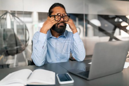 Photo for Tired young indian man in formal outwear employee suffering from sore eyes, sitting at desk in front of laptop at office, rubbing his eyes under eyeglasses, copy space - Royalty Free Image