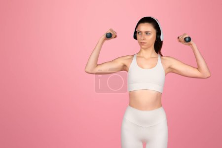 Téléchargez les photos : Focalisé grave caucasienne jeune femme en blanc athlétique porter des muscles fléchissants avec des haltères tout en portant un casque sur un fond rose. Bien-être, musique, soin du corps - en image libre de droit