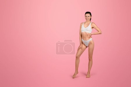 Foto de Una joven caucásica confiada en un sujetador deportivo blanco y calzoncillos a juego golpea una pose juguetona sobre un fondo rosado, irradiando salud y forma física. Bienestar, estilo de vida corporal - Imagen libre de derechos