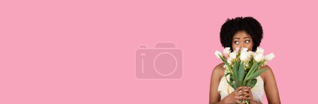 Téléchargez les photos : Jeune femme afro-américaine exubérante aux cheveux bouclés et à l'expression joyeuse, tenant un bouquet de tulipes blanches et roses, évoquant un sentiment de fraîcheur, de printemps et de bonheur sur un fond rose vibrant - en image libre de droit