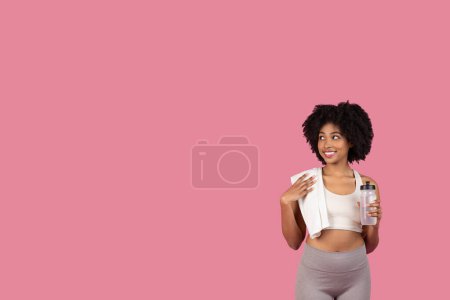 Photo for Cheerful athletic black woman holding water bottle and draping towel over her shoulder, taking break from her fitness routine against pink background, looking at free space - Royalty Free Image