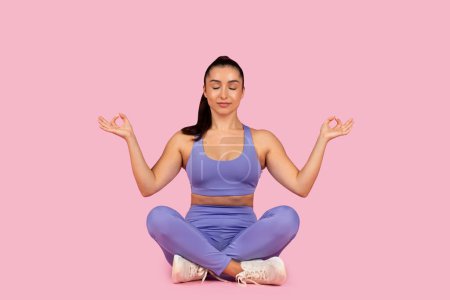 Photo for Serenely poised woman in meditative yoga pose, dressed in purple activewear, sits with eyes closed against tranquil pink background, symbolizing peace - Royalty Free Image