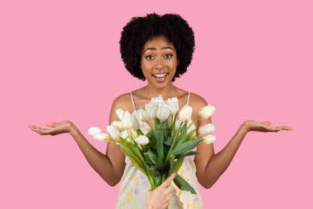 Photo for Surprised and happy African American millennial woman presenting a lush bouquet of white tulips, with a playful expression, against a vibrant pink background, studio. People emotions - Royalty Free Image