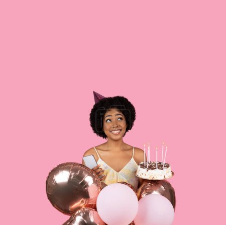 Téléchargez les photos : Joyeuse jeune femme afro-américaine aux cheveux bouclés naturels, portant un chapeau d'anniversaire, tenant des ballons et un gâteau aux bougies, prête à célébrer, sur fond rose avec amplement d'espace de copie - en image libre de droit