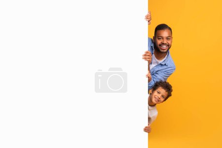 Téléchargez les photos : Père et fils noirs regardant hors du panneau publicitaire vierge, homme afro-américain joyeux et son enfant mâle debout près du grand panneau blanc, démontrant l'espace libre de copie pour votre conception ou offre - en image libre de droit