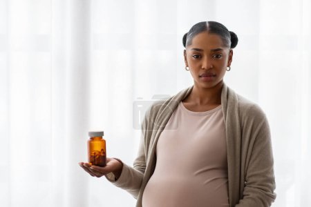 Serious young pregnant black woman wearing casual outfit showing jar with medicine, unwilling to take multivitamins, supplements during pregnancy, blank copy space