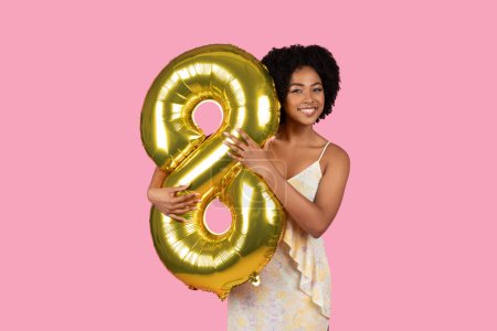 Foto de Radiante joven afroamericana con el pelo rizado sosteniendo un globo número 8 de oro, celebrando una ocasión especial, vacaciones de primavera con alegría y entusiasmo en un telón de fondo rosa - Imagen libre de derechos