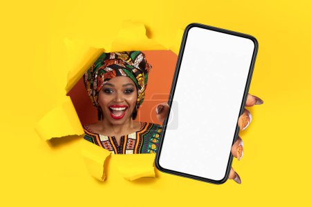 Femme afro-américaine dynamique en tenue traditionnelle avec le sourire regardant à travers un papier jaune déchiré, tenant un smartphone avec un écran vide. Grande offre, annonce, site de recommandation, application
