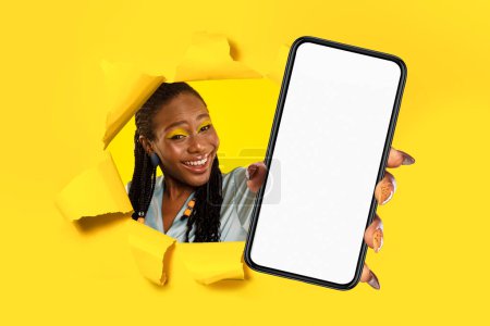 Bright and cheerful african american woman with braids and colorful makeup peeks through a yellow torn background, holding a smartphone with a blank screen, ideal for app demos