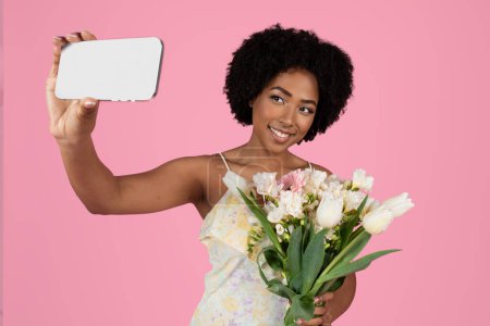 Photo for Charming smiling young african american woman with curly hair taking a selfie, holding a smartphone and a bouquet of white and pink tulips on a soft pink background, studio - Royalty Free Image