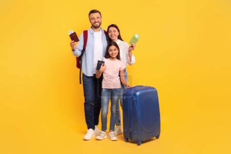 Téléchargez les photos : Joyeuse famille de trois personnes prête pour le vol montrant passeports et billets, souriant à la caméra rêvant de la prochaine aventure de vacances, debout avec valise sur fond de studio jaune, pleine longueur - en image libre de droit