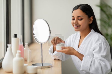 Téléchargez les photos : Portrait d'une femme indienne attrayante appliquant de la crème pour le visage, posant avec de l'hydratant dans ses mains, assis dans la salle de bain moderne à l'intérieur. Produits cosmétiques pour la peau Concept - en image libre de droit
