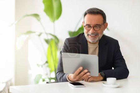 Téléchargez les photos : Homme mûr caucasien joyeux en costume intelligent et col roulé, s'engageant avec une tablette numérique, assis dans un espace de bureau bien éclairé avec des plantes vertes et une tasse à café. Travail, entreprise - en image libre de droit