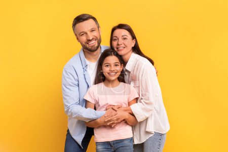 Photo for Young family of three shares happy moment with warm hugs and smiles, standing together in studio with yellow backdrop. Portrait of cheerful parents and preteen daughter bonding and embracing - Royalty Free Image