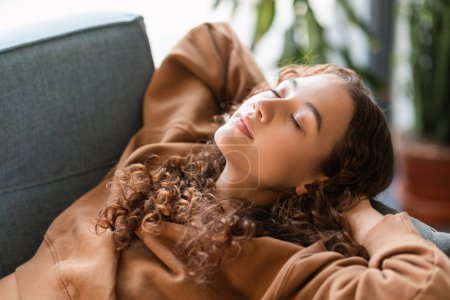 Photo for College student girl enjoys peaceful nap on comfortable sofa indoors, resting with closed eyes in deserved relaxation. Female teenager napping holding hands behind head, closeup - Royalty Free Image