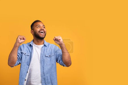 Foto de Gran Suerte Concepto. Hombre negro alegre celebrando el éxito con los brazos levantados, chico afroamericano feliz sacudiendo puños y exclamando con emoción, de pie aislado en el fondo del estudio amarillo - Imagen libre de derechos