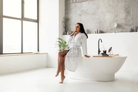 Téléchargez les photos : Joyeux attrayant jeune femme orientale en peignoir blanc assis sur la baignoire et regardant par la fenêtre panoramique à la salle de bain de luxe, pleine longueur, espace de copie. Home spa - en image libre de droit