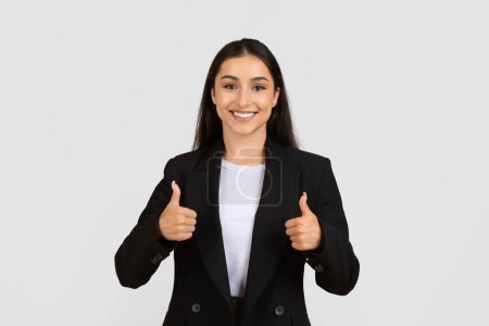 Photo for Cheerful young businesswoman with beaming smile, dressed in formal black suit, giving two thumbs up, signifying approval and success, isolated on light grey background - Royalty Free Image