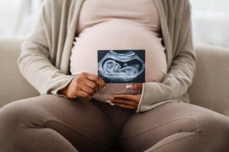 Photo for Unrecognizable african american expecting woman with big belly sitting on couch at home, holding sonogram ultrasound image of her baby and hugging tummy, cropped - Royalty Free Image