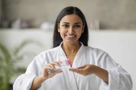 Téléchargez les photos : Jeune femme indienne positive portant un peignoir blanc hydratant visage dans la salle de bain, en utilisant du coton tampon et toner, appliquer du maquillage, regarder la caméra et sourire - en image libre de droit