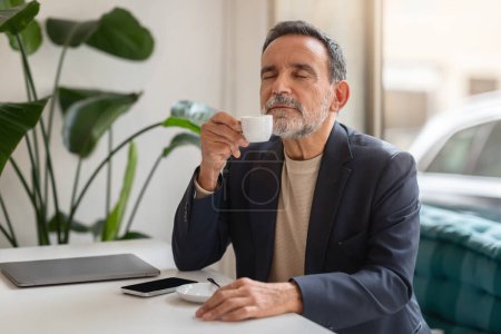 Content happy caucasian senior businessman in a dark blazer savoring a cup of espresso with closed eyes, sitting at a cafe table with a laptop and smartphone, enjoy spare time