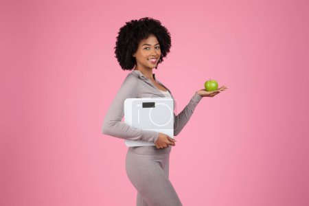 Foto de Sonriente joven mujer negra balanceando manzana verde en una mano y sosteniendo balanza en la otra, promoviendo dieta saludable y control de peso sobre fondo rosa - Imagen libre de derechos