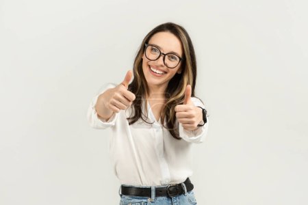 Téléchargez les photos : Jeune femme européenne enthousiaste avec des cheveux bruns et des lunettes donnant deux pouces vers le haut, portant un chemisier blanc et un jean, exsudant positivité et confiance sur un fond clair - en image libre de droit