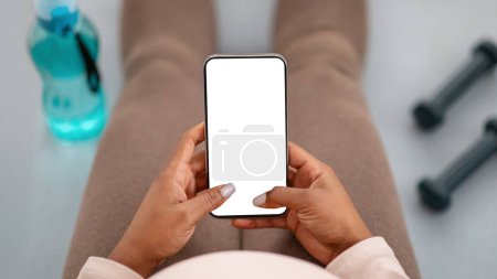 Foto de Cultivado de mujer embarazada deportiva afroamericana sentada en una esterilla de yoga durante el entrenamiento con pesas, utilizando un teléfono inteligente con la maqueta de pantalla blanca en blanco, comprobando la aplicación móvil de fitness para futuras madres - Imagen libre de derechos