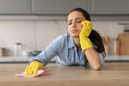 Foto de Mujer joven cansada con guantes amarillos apoyando su cabeza en su mano mientras limpia la encimera de la cocina con tela rosa, sintiéndose agotada, espacio libre - Imagen libre de derechos