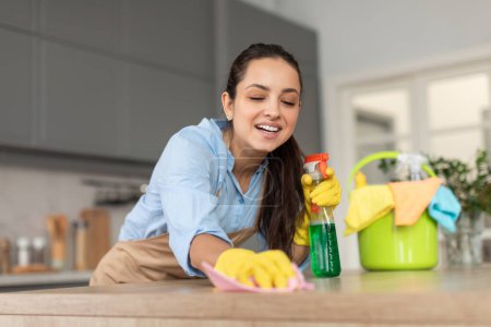 Foto de Mujer sonriente en ropa casual y guantes de goma limpiando con entusiasmo la encimera de la cocina con botella de aerosol y tela, con cubo cerca - Imagen libre de derechos