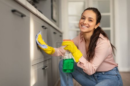 Téléchargez les photos : Femme souriante en chemise rose et gants jaunes est accroupi tout en nettoyant les portes de l'armoire de cuisine avec un chiffon bleu et une bouteille de pulvérisation, copiez l'espace - en image libre de droit