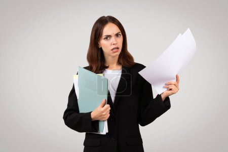 Photo for A puzzled millennial european businesswoman in a suit frowns while holding documents and a folder, depicting confusion or concern in a corporate setting, isolated on a gray background - Royalty Free Image