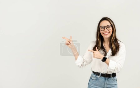 Photo for Happy european woman with glasses pointing fingers to the side, wearing a white shirt and jeans, with a smartwatch and a cheerful expression on a light background, studio - Royalty Free Image