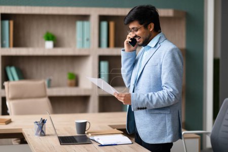 Téléchargez les photos : Un homme d'affaires indien prospère discute de documents au téléphone à l'intérieur du bureau, regarde des documents et parle sur son téléphone portable au travail, examine des documents et discute des détails - en image libre de droit