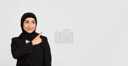 Photo for Pleasant woman in black hijab and suit jacket gesturing to the right at free space with confident smile, standing against spacious grey backdrop, banner - Royalty Free Image