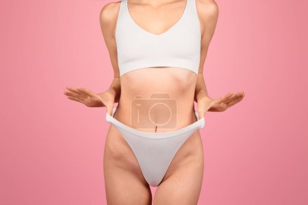 Foto de Una vista de sección media de la mujer caucásica en ropa deportiva blanca, las manos señalando hacia su abdomen en forma, haciendo hincapié en un cuerpo sano sobre un telón de fondo rosa. Bienestar, resultado del cuidado corporal - Imagen libre de derechos
