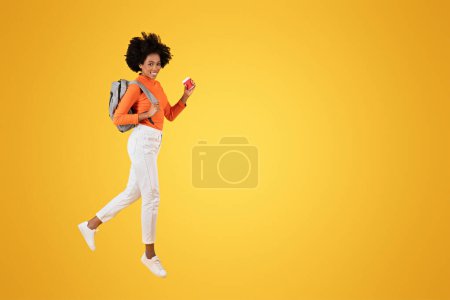 Foto de Mujer afroamericana enérgica con el pelo rizado, con una camisa naranja, jeans blancos y zapatillas de deporte, saltando felizmente con una mochila gris y sosteniendo una taza roja sobre un fondo amarillo brillante - Imagen libre de derechos