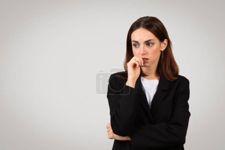 Téléchargez les photos : Femme d'affaires européenne millénaire soucieuse de se mordre les ongles alors qu'elle est profondément dans la pensée, affichant anxiété ou nervosité, avec un regard coûteux, dans un costume d'affaires noir classique - en image libre de droit