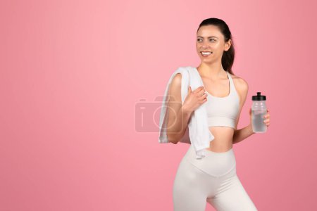 Active and healthy european young woman in white athletic wear with a towel over her shoulder and a water bottle in hand, looking refreshed after a workout on a pink background