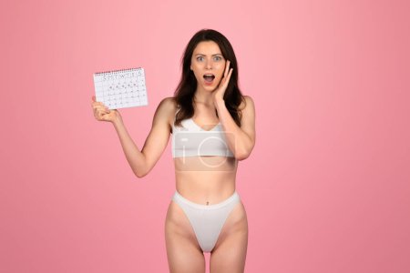 Photo for Shocked glad young caucasian woman in white underwear holding a calendar with a surprised expression, symbolizing forgotten events or deadlines on a pink background. Wellness, body care lifestyle - Royalty Free Image